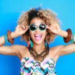 Black woman against a blue wall smiles while wearing sunglasses and summer clothes after her summer smile makeover