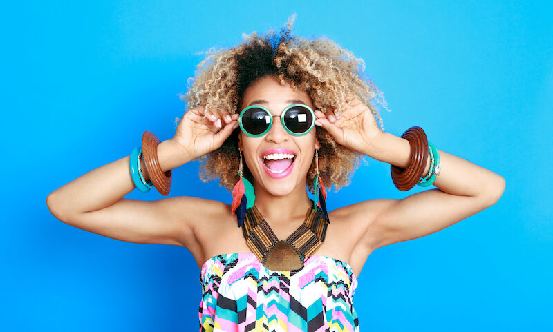 Black woman against a blue wall smiles while wearing sunglasses and summer clothes after her summer smile makeover