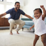 Black date crouches in the living room and smiles at his baby girl as she walks happily