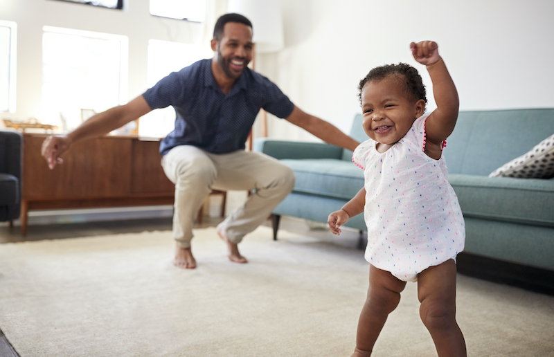 Black date crouches in the living room and smiles at his baby girl as she walks happily