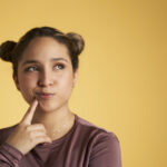 Young woman against a yellow background touches her chin and looks up wondering if Six Month Smiles or clear aligners are right for her smile