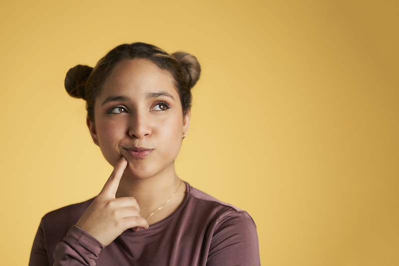 Young woman against a yellow background touches her chin and looks up wondering if Six Month Smiles or clear aligners are right for her smile