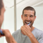 Brunette man in a gray shirt smiles in his bathroom mirror while brushing his teeth