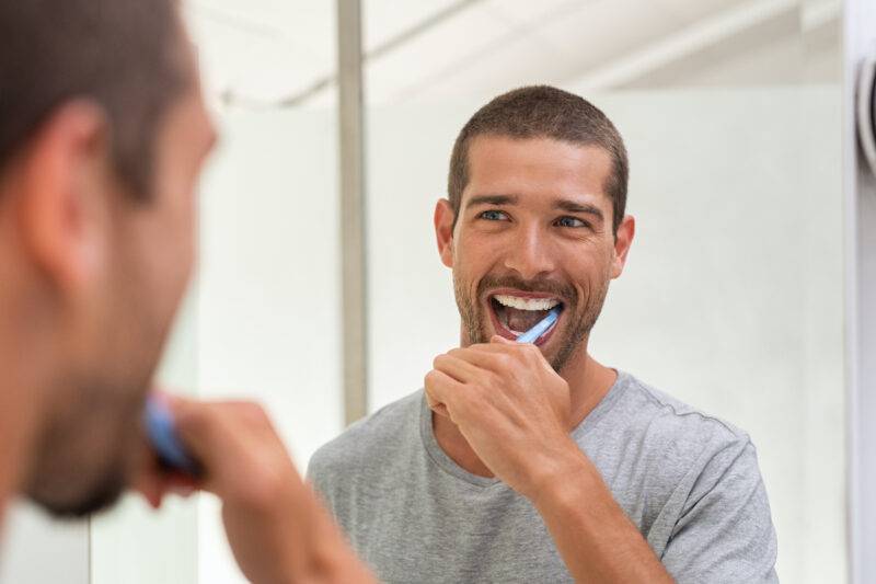 Brunette man in a gray shirt smiles in his bathroom mirror while brushing his teeth